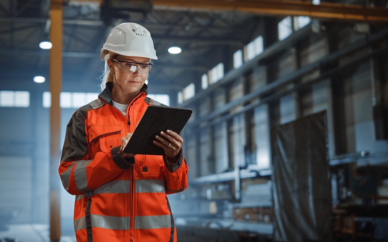 Image of a woman in safety gear and a fluorescent orange jacket working on a tablet in a warehouse